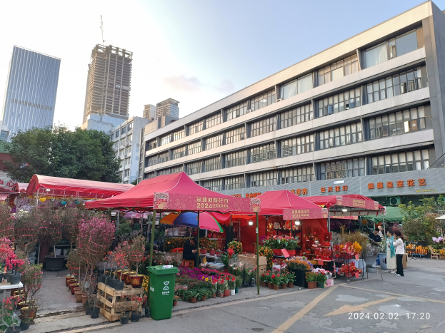 Ein blühender Blumenmarkt mit bunten Blumen, Pflanzen und Bäumen zwischen hohen Gebäuden in einer Stadtstraße, mit Menschen, die herumlaufen und weißen Wolken am Himmel.
