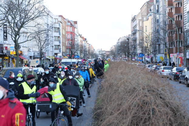 Eine große Gruppe von Menschen in Masken und Sicherheitswesten auf Fahrrädern auf einer von Bäumen gesäumten Straße mit Fahrzeugen, Laternenpfählen und Texttafeln unter einem klaren blauen Himmel.
