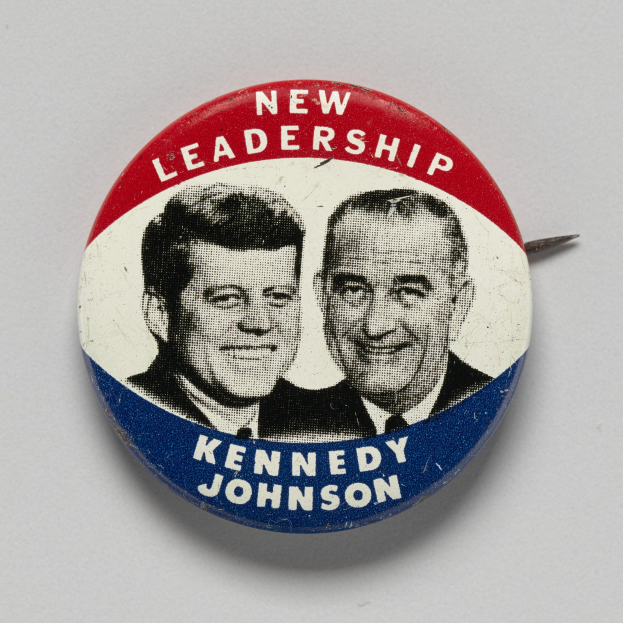 Ein Ansteckbutton auf einer weißen Oberfläche zeigt zwei Bilder von Kennedy und Johnson mit dem Text "Neue Führung" darunter.