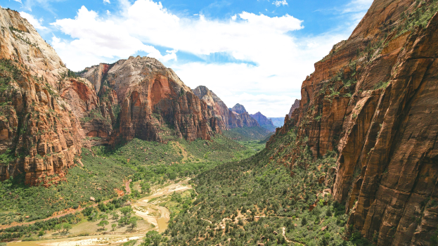 Atemberaubender Blick auf den Zion-Nationalpark in Utah mit saftig grünen Bäumen, sanften Hügeln, einem ruhigen Gewässer im Vordergrund und einem Himmel mit flauschigen weißen Wolken.