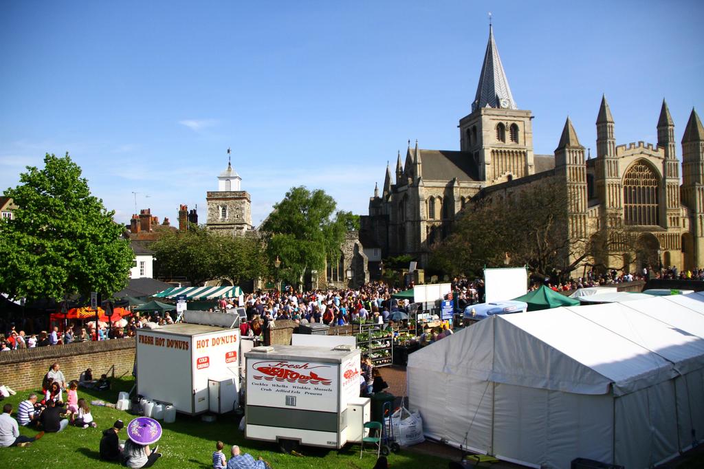 Historische Gebäude mit Bäumen davor, zusätzliche Gebäude und ein Turm im Hintergrund, ein Weg mit Menschen und Geschäften und Individuen, die auf dem Rasen in der Nähe der Geschäfte sitzen.