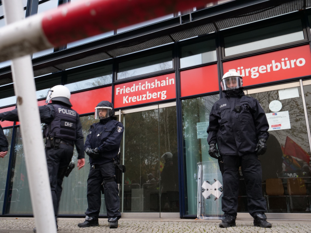 Polizeibeamte in Uniform stehen vor einem Glasgebäude mit einer Stange auf der linken Seite und Schildern mit der Aufschrift 'Friedrichshain Kreuzberg' im Hintergrund.