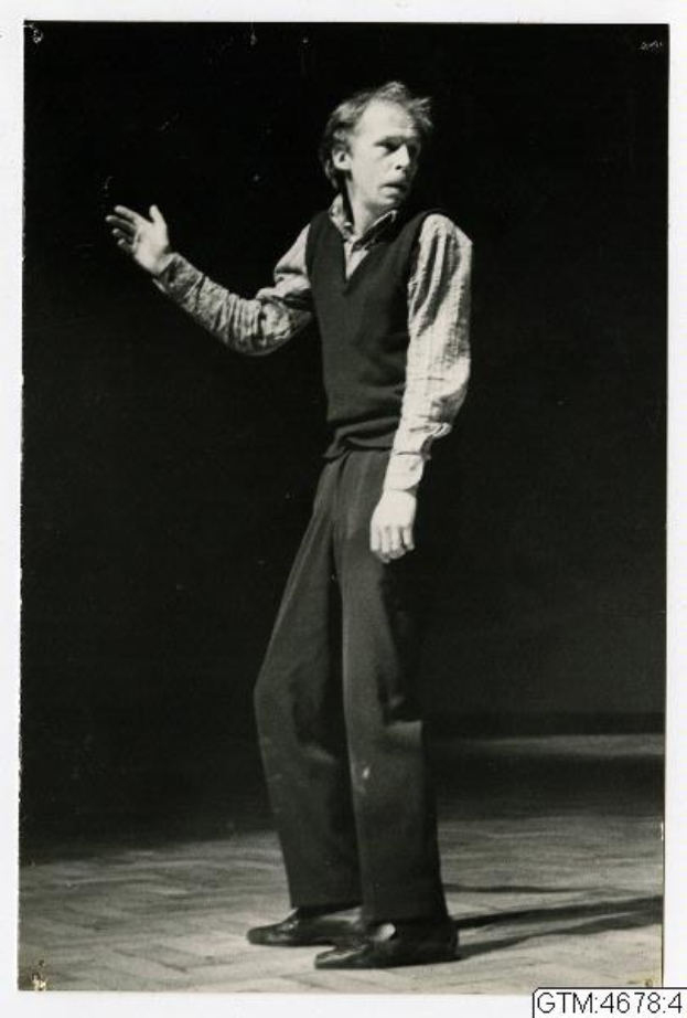 Ein Mann in einem weißen Hemd und dunklen Hosen steht auf einer Bühne mit ausgestreckten Armen, geneigtem Kopf und ernstem Gesichtsausdruck.