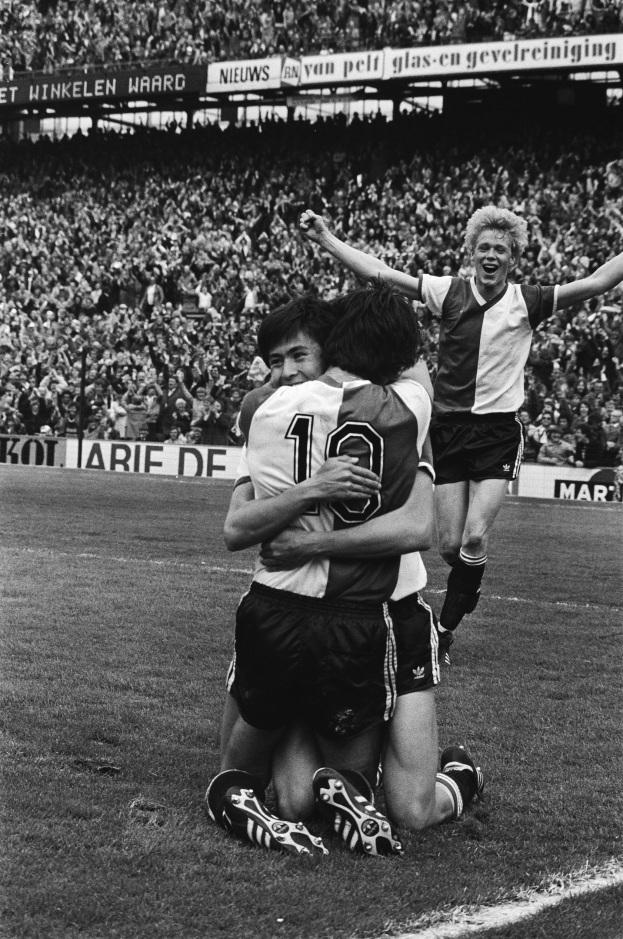 Drei Füßballspieler umarmen sich auf dem Boden nach einem Tor, mit jubelnder Menge und schwenkenden Fahnen in einem schwarz-weiß gestalteten Stadion.