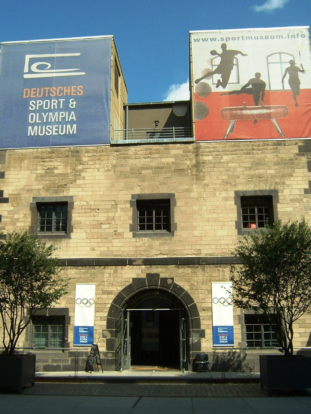 Außenansicht des Deutschen Sport & Olympia Museums in Berlin mit einem Gebäude mit Fenstern und einer Tür, umgeben von Bäumen und Bannern, unter einem bewölkten Himmel.