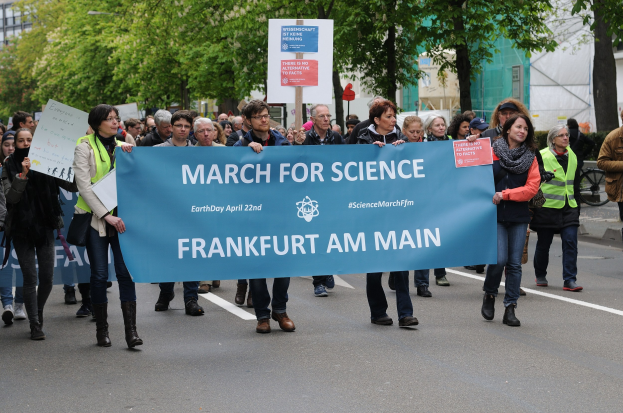 Eine Gruppe von Menschen marschiert mit einem "March for Science Frankfurt am Main"-Schild die Straße entlang, mit Bäumen und Gebäuden im Hintergrund.