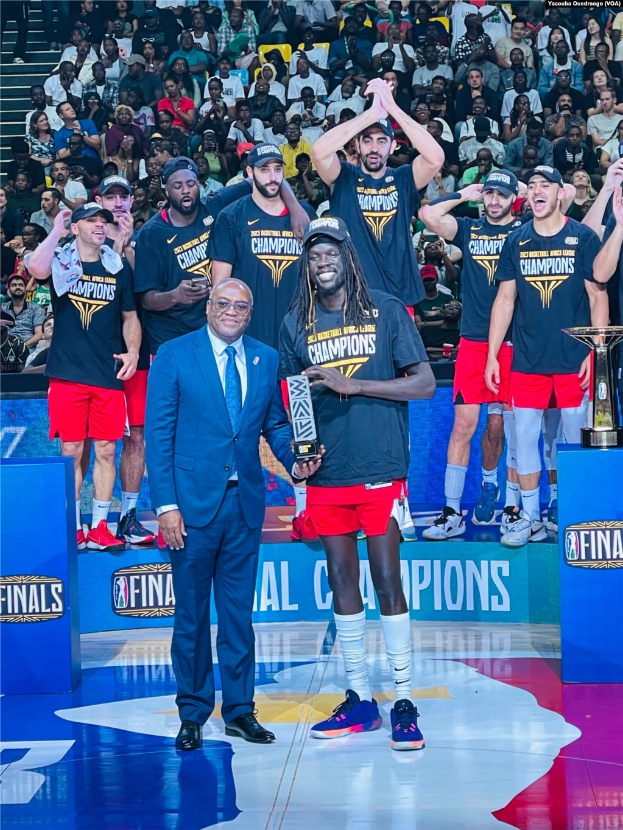 Eine Gruppe von Männern auf einem Basketballfeld feiert, zwei halten Trophäen, mit Zuschauern im Hintergrund und zwei Trophäen auf Podesten rechts.
