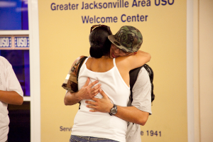 Eine Frau in einem weißen T-Shirt umarmt einen Mann in einem weißen T-Shirt, einer Brille und einer Kappe, mit einer Handtasche über der Schulter, vor einem gelben und blauen Schild mit Text.