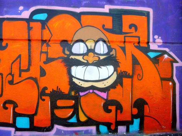 Ein lebendiges Graffiti-Gemälde eines Cartoon-Charakters mit großen Augen, einem breiten Grinsen und einem verschmitzten Ausdruck an der Seite eines Gebäudes, begleitet von auffälligem Text in einem fröhlichen, bunten Stil.