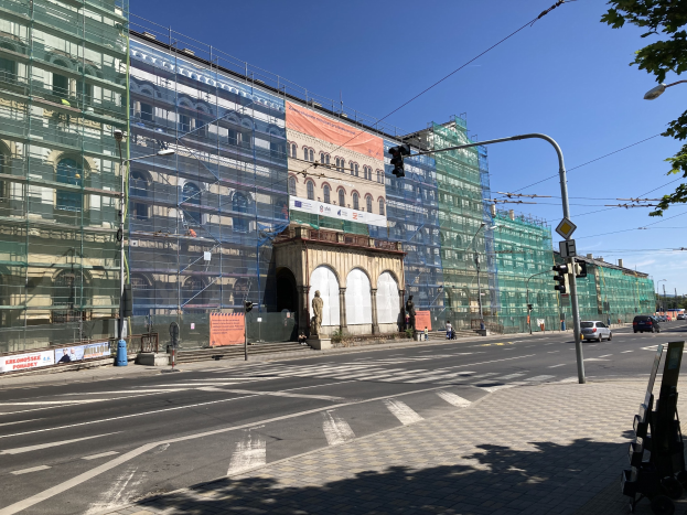 Großer mehrgeschossiger Bau unter construction in Berlin, umgeben von Straßeninfrastruktur und Fahrzeugen, mit Fußgängern auf dem Gehweg und Bäumen im Hintergrund.