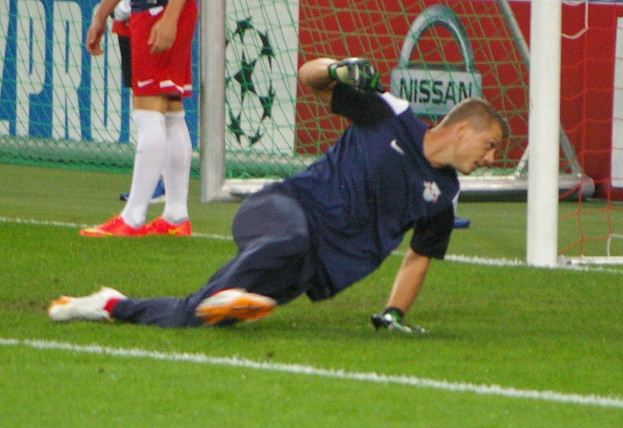 Ein Fußballer mit Handschuhen taucht auf, um einen Ball vor einem Tor zu fangen, mit zwei Personen, einem Netz und einer Banner mit Text im Hintergrund.