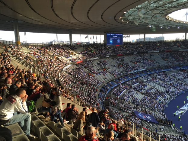 Eine große Menschenmenge sitzt im Allianz Stadion in München, Deutschland, und schaut ein Fußballspiel. Auf der rechten Seite befindet sich eine Bühne mit ein paar Leuten darauf und im Hintergrund sind Fahnen, Stangen und ein Bildschirm zu sehen.
