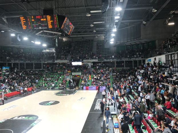 Eine große Halle mit Zuschauern, die ein Basketballspiel verfolgen, mit einem Basketballkorb links, Anzeigetafeln, Bildschirmen und Deckenbeleuchtung im Hintergrund.