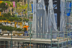 Eine Baustelle mit Gerüsten an einem Gebäude, mit verschiedenen Objekten und Personen im Vordergrund und Gebäuden und Bäumen im Hintergrund, mit einem Wasserzeichen unten.