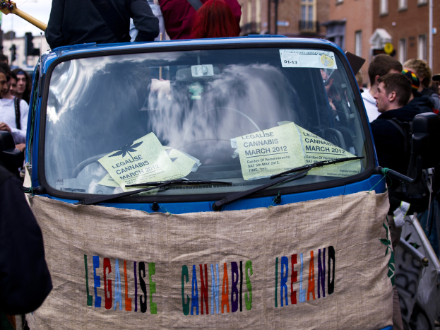 Eine Gruppe von Menschen steht um ein Auto mit einem "Legalise Cannabis Ireland"-Schild herum, umgeben von Gebäuden mit Fenstern und Laternen unter einem klaren blauen Himmel, mit Papieren im Auto sichtbar.