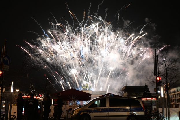 Polizeiauto vor Menschenmenge bei Silvesterfeier in Berlin geparkt, mit farbigen Feuerwerken am Himmel.