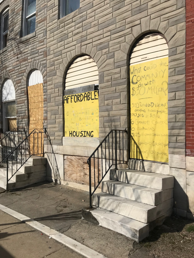 Verschlossenes Gebäude mit "Bezahlbarer Wohnraum"-Schild, Treppe mit Geländer und einer Straße unten.