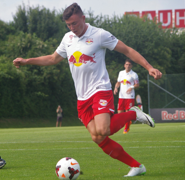 Ein Mann in einem Fußballtrikot schießt einen Ball auf einem grünen Feld mit Bäumen und einem klaren blauen Himmel im Hintergrund, wobei ein Schild mit der Aufschrift "RB Leipzig" sichtbar ist.