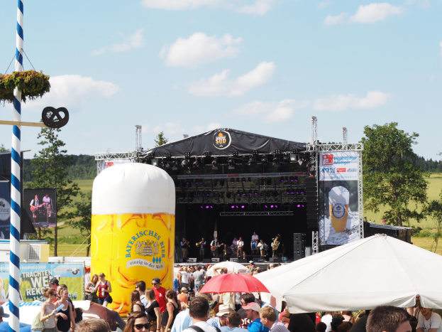 Eine große Menschenmenge beim Oktoberfest in München, Deutschland, viele tragen Mützen und Goggles, einige halten Schirme, mit einem Zelt im Vordergrund, einem dekorierten Pfahl links und einer Bühne im Hintergrund mit einem Banner und Musikern.