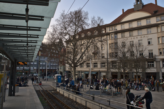 Eine belebte Straßenkreuzung mit einem Bahnhof auf der linken Seite, umgeben von Gebäuden, Bäumen, Laternen und Textanzeigen, wo Menschen gehen, auf Bänken sitzen oder stehen, unter einem sichtbaren Himmel.