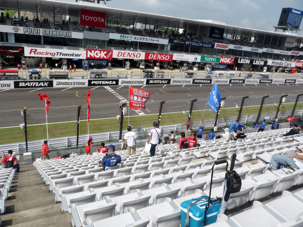 Eine Gruppe von Menschen sitzt auf Tribünenplätzen einer Rennstrecke mit Fahnen, Texttafeln, Geländern und Stufen unter einem bewölkten Himmel.
