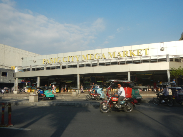 Eine belebte Szene am Pasig City Mega Market mit Fahrzeugen auf der Straße, Fußgängern, einem Gebäude mit Text im Hintergrund und einem bewölkten Himmel.