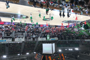 Eine Gruppe von Menschen spielt Basketball in einer großen Arena, mit Zuschauern auf den Tribünen und beleuchtet von Bildschirmen und Lichtern am unteren Bildrand.