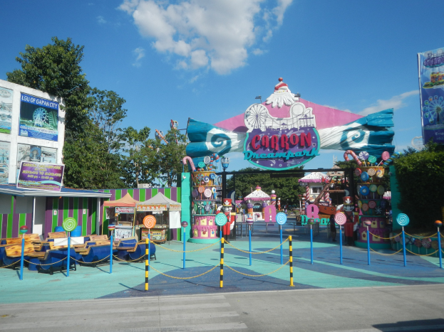 Kirmesfahrgeschäft am Parkeingang mit Straße darunter, eingezäuntes Gebiet, Buden, Banner, Bäume und bewölkter Himmel.