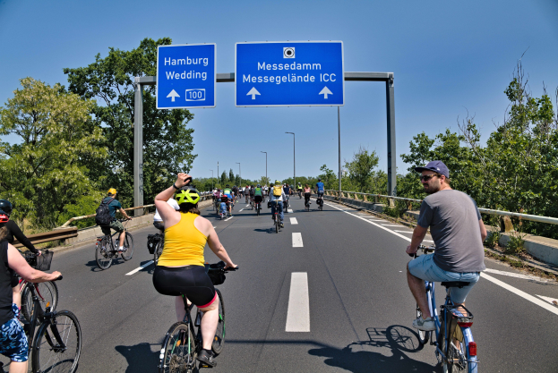Gruppe von Radfahrern in Helmen auf einer Straße mit einem Geländer und Bäumen auf beiden Seiten, Laternen im Hintergrund und einem klaren blauen Himmel, mit einem Schild, das eine Radtour in Hamburg anzeigt.
