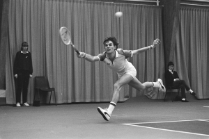 Ein Mann schwingt einen Tennis-Sch├Ąger gegen einen Ball, mit zwei stehenden und zwei sitzenden Personen im Hintergrund und einem Vorhang als Rahmen.