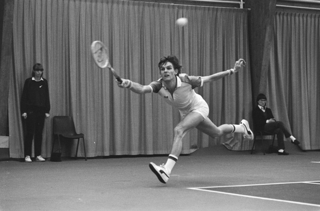Ein Mann schwingt einen Tennis-Sch├Ąger gegen einen Ball, mit zwei stehenden und zwei sitzenden Personen im Hintergrund und einem Vorhang als Rahmen.