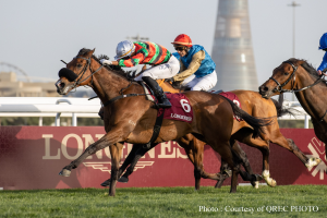 Drei Jockeys auf Pferden, die auf einer saftig grünen Rennbahn unter einem klaren blauen Himmel galoppieren, mit einem fernen Turm und einer Tafel mit Text im Hintergrund.