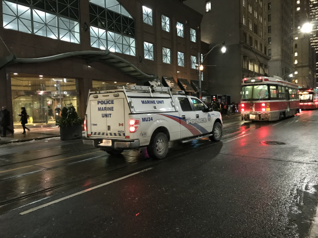 Ein Polizei-Lkw steht nachts am Straßenrand, mit Fahrzeugen auf der Straße, Fußgängern auf dem Gehweg, Topfpflanzen, Straßenlaternen, Gebäuden mit Fenstern und einer bewölkten Himmel.
