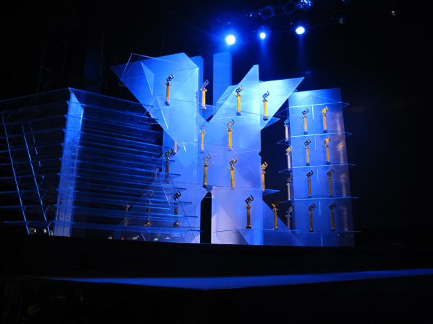 Eine Bühne für eine Preisverleihung mit dunklem Hintergrund, beleuchtet von ein paar Lichtern und mit mehreren Gegenständen, die mit dem Ereignis in Verbindung stehen.