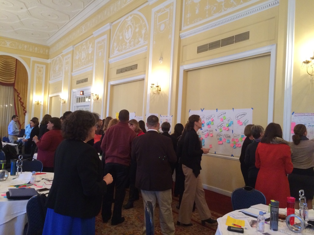 Eine Gruppe von Menschen untersucht Plakate und Charts, die an einer Wand in einem scheinbar künstlerischen Museum angezeigt werden.