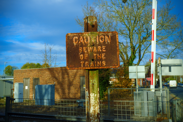 Warnschild an einer Bahnschiene umgeben von einem Metallzaun, Bäumen, Strommasten, einem Gebäude mit Fenstern, Containern, einer Straßenlaterne, Fahrzeugen auf der Straße und einem bewölkten Himmel.