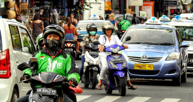 Eine Gruppe von Motorradfahrern fährt eine belebte Straße entlang, neben Autos und anderen Fahrzeugen, wobei einige Fahrer Helme tragen, Gebäude, Bäume und Geschäfte sind im Hintergrund zu sehen.