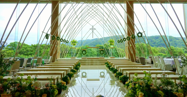 Eine Hochzeitszeremonie in einem großen Saal mit Tischen in Halbkreisform, geschmückt mit Blumenbouquets, unter einem klaren blauen Himmel mit Bäumen und Hügeln im Hintergrund.