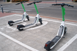 Drei weiße und grüne Elektro-Scooter nebeneinander auf einer Straße mit Pfählen, Pflanzen, Bäumen, Gebäuden und Himmel im Hintergrund.