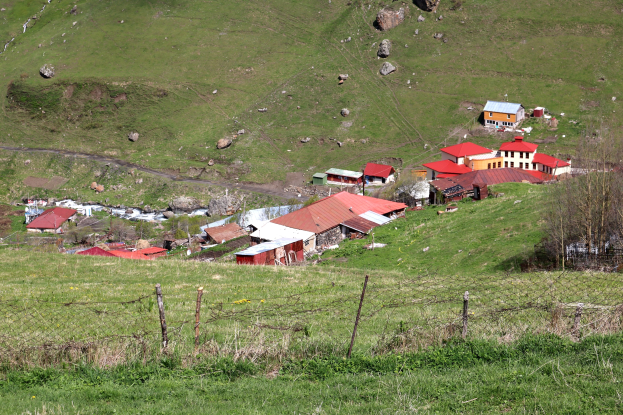 Kleines Dorf umgeben von einer Einfriedung, eingebettet in eine grüne Hügellandschaft mit verstreuten Felsen und Grasland.
