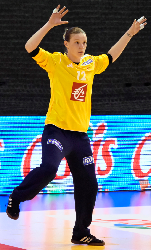 Eine Frau in einem gelben T-Shirt und schwarzen Hosen, die Handball spielt, mit einem Bildschirm im Hintergrund.