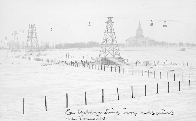 Ein Schwarz-Weiß-Foto einer Skiliftanlage in einer verschneiten Landschaft, umgeben von Pfählen, Seilen, Bäumen und einem Gebäude im Hintergrund, mit Text am unteren Rand.