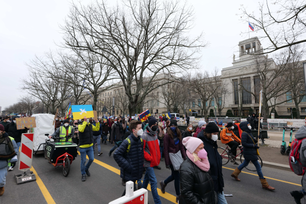 Eine große Gruppe von Menschen marschiert bei einer Demonstration die Straße in Washington, D.C. entlang, mit Schildern und Bannern, einige auf Fahrrädern, unter einem klaren blauen Himmel vor einem Gebäude.