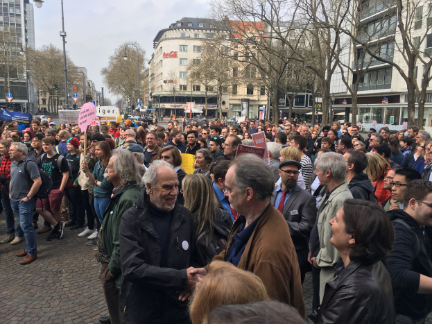 Große Menschenmenge bei einer Demonstration in Berlin, Deutschland, mit Schildern und in der Nähe von Fahrzeugen, Bäumen und Gebäuden mit Fenstern.