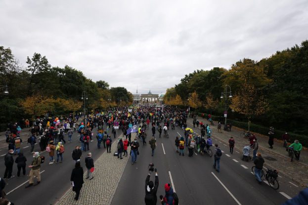 Eine große Gruppe von Menschen marschiert auf einer baumbestandenen Straße in Berlin, mit Kameras in der Hand, mit einem Gebäude und einem klaren Himmel im Hintergrund.