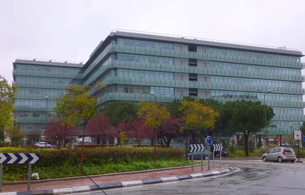 Großes modernes Gebäude mit zahlreichen Fenstern, als Europäische Zentralbank-Hauptquartier identifiziert, umgeben von Grünflächen, Straßeninfrastruktur und Fahrzeugen unter einem bewölkten Himmel.