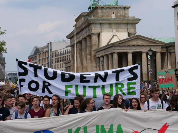 Gruppe von Schülern marschiert in Berlin mit einem bunt bemalten 'Students for Future' Banner vor Gebäuden, Bäumen und Himmel.