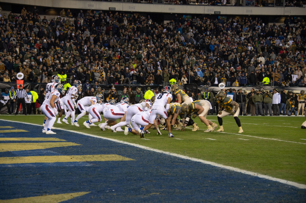 Ein Fußballspiel auf einem Stadionfeld mit Spielern in Sportkleidung und Helmen auf dem Boden, umgeben von Zuschauern und Stadionmerkmalen.
