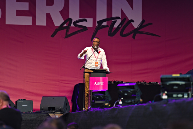 Ein Mann in einem weißen Hemd und braunen Hosen steht auf einer Bühne und hält ein Mikrofon in der Hand, während ein pinkfarbenes Banner mit der Aufschrift 'Berlin as Fuck' hinter ihm und Lautsprecher auf der Bühne zu sehen sind.