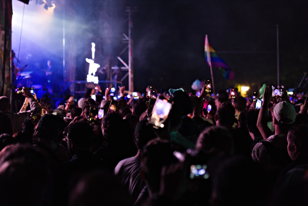 Menge bei einem Konzert mit hochgehaltenen Handys, beleuchtet von Lichtern, umgeben von Pfählen und einer Flagge, während Musiker auf der Bühne spielen.